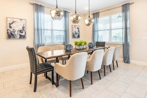 A dining room with a wooden table, six chairs, two hanging pendant lights, and a window with blinds.