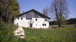 Un bâtiment blanc avec un toit en pente, un chemin de pierre menant à l’entrée, et un champ verdoyant parsemé d’arbres, sous un ciel dégagé.