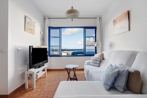 Una habitación con televisión sobre un mueble bajo, un sofá, una mesita y una ventana con vista al mar.