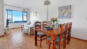Un comedor con una mesa de madera, sillas y un jarrón de flores. Se ve una ventana con vistas al mar.