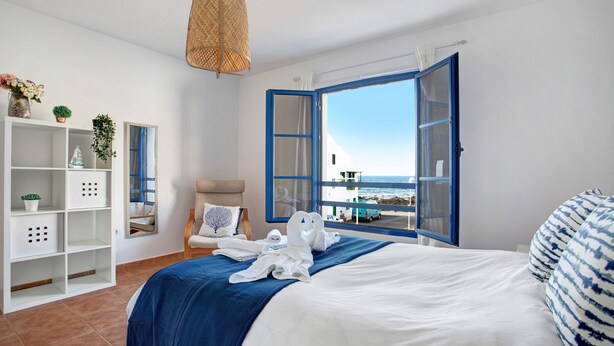 Habitación de hotel con cama, un armario blanco, una silla, ventana con persianas azules y vista al mar.