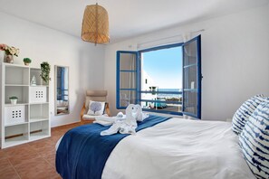 Una habitació d'hotel amb un llit, un armari blanc, una cadira, una finestra amb persianes blaves i vista al mar.