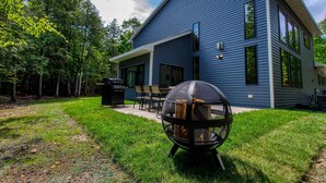 A modern two-story house with a covered patio, a fire pit, and a view of a forested area. - Expansive Modern Loft House Sleeps 12 (Egg Harbor)