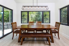 Une salle à manger avec une grande table en bois, quatre chaises, un banc et de grandes fenêtres donnant sur la forêt.