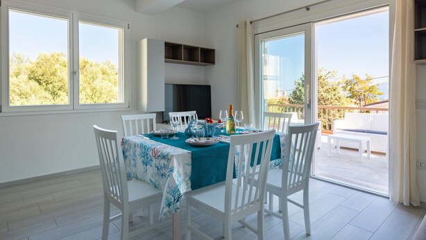 A dining area with a table set for four, white chairs, a teal tablecloth, and a view of the outdoors through large windows.