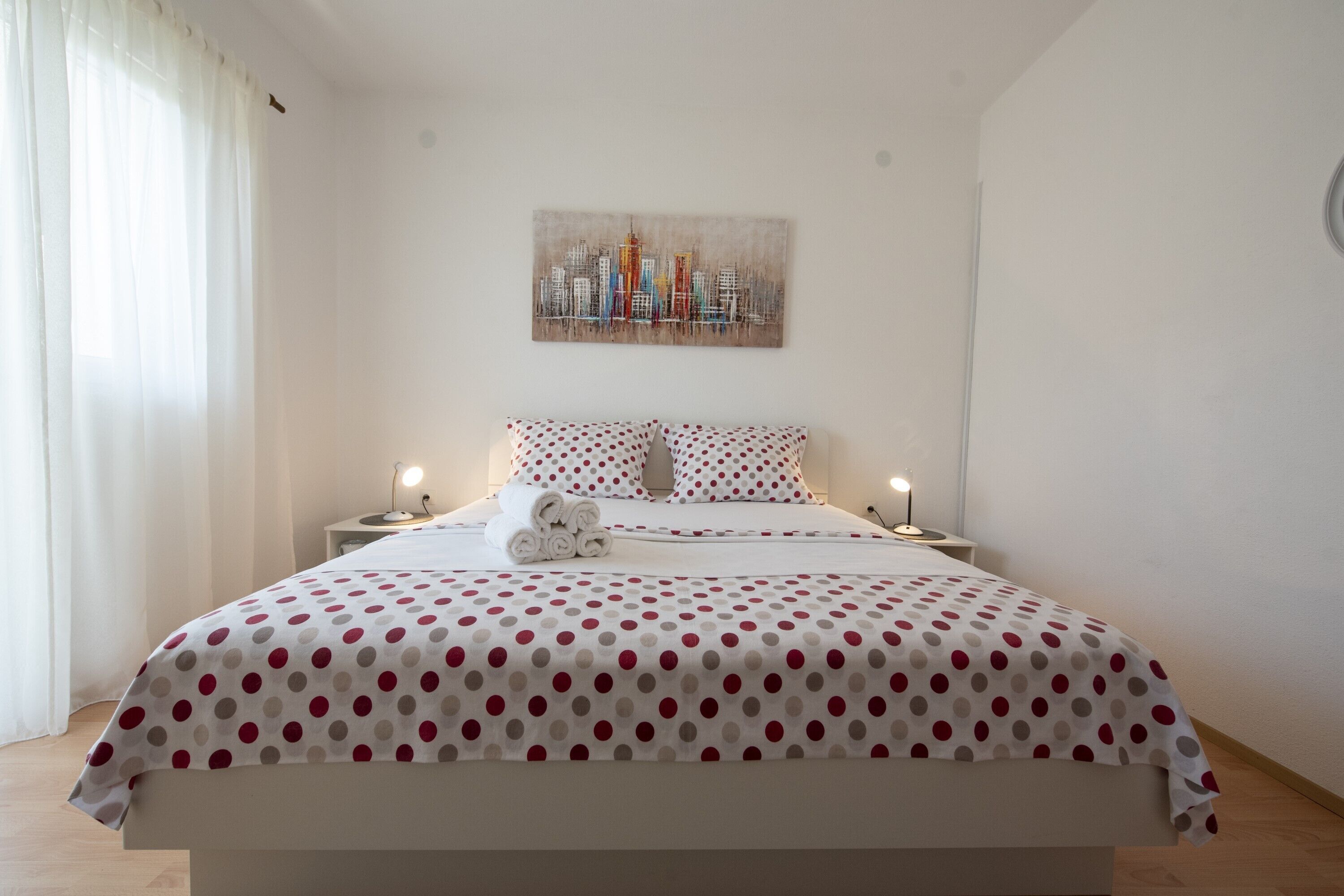 A neatly made bed with a polka dot bedspread, a cityscape painting above it, and two bedside lamps.