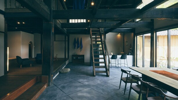 Una habitación amplia con techo alto, una escalera de madera, un comedor con una larga mesa y sillas, y vistas a través de grandes ventanales.