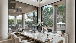 Une cuisine moderne dotée d’un grand îlot central, de tabourets de bar et offrant une vue sur l’espace extérieur, agrémenté de parasols et de verdure.