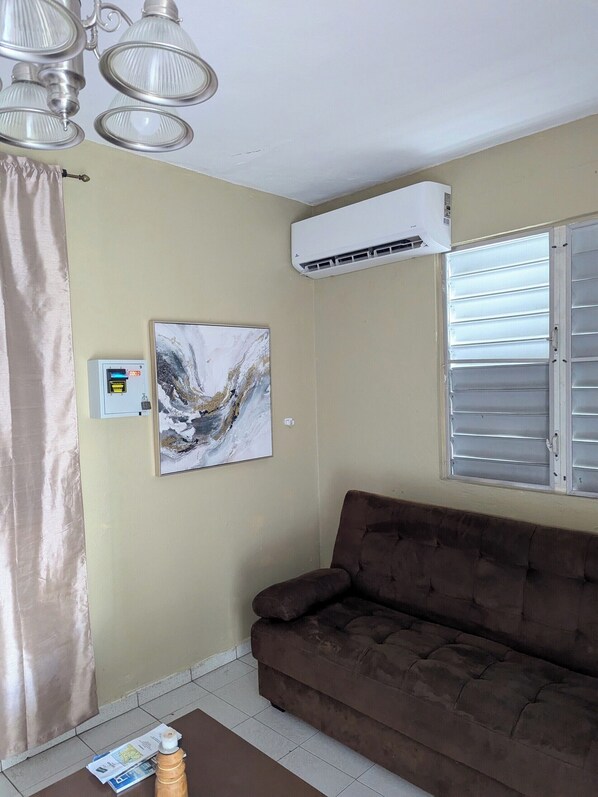 Ein Wohnzimmer mit einem braunen Sofa, einer Deckenleuchte, einer Klimaanlage, einem Fenster mit Jalousien und einem an der Wand befestigten Luftreiniger.