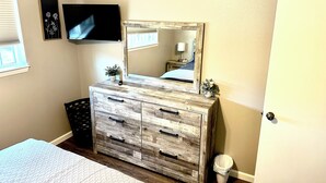 A bedroom with a wooden dresser, a television mounted on the wall, a mirror, and a small trash bin.