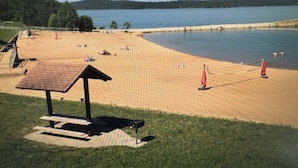 A beach with a picnic table and umbrellas, a lake in the background, and a grassy area in the foreground. - Mercy's Hideaway - Open Pool! Relaxing & Chic! Prime Location w/ Natural Views! (Branson)