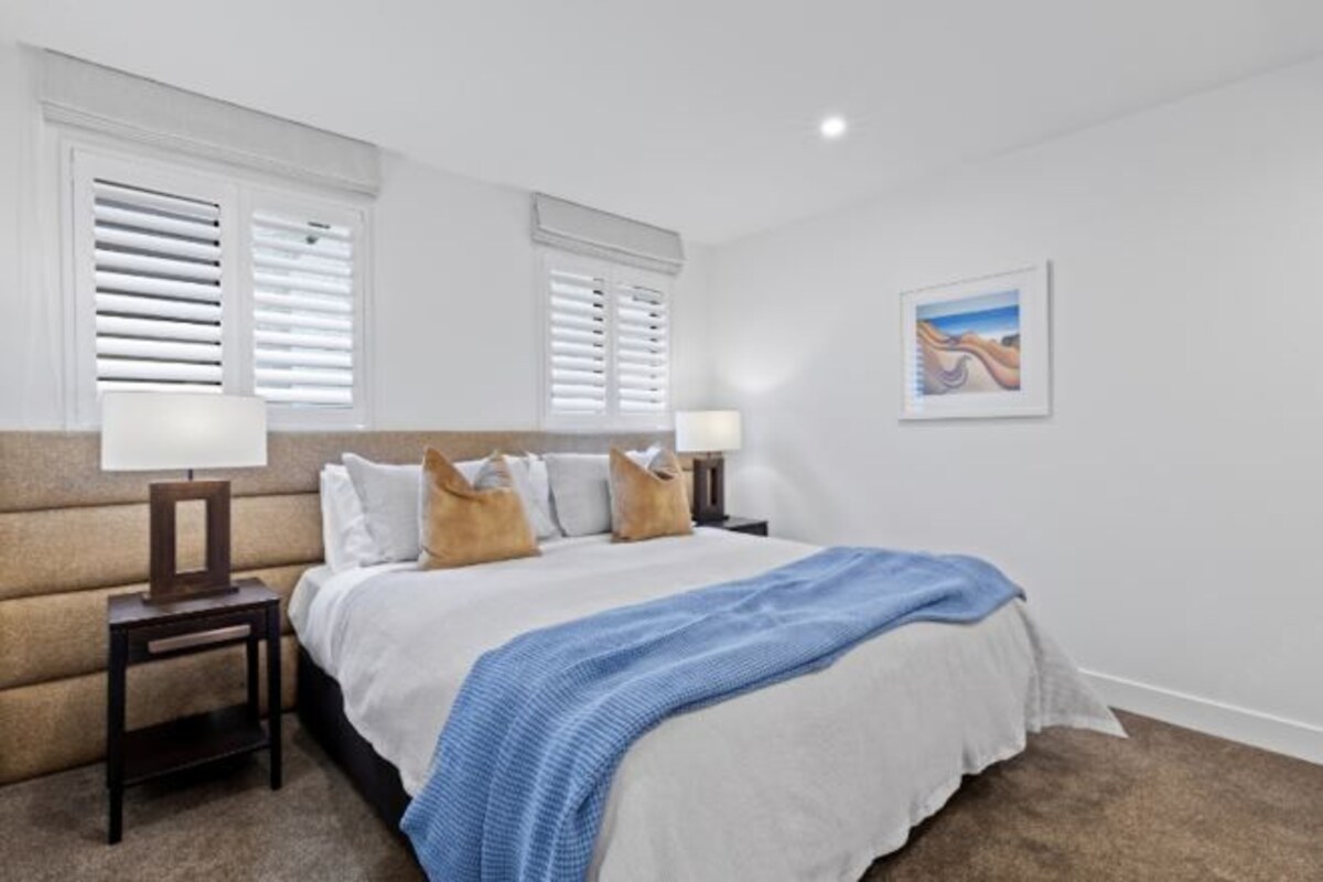 A bedroom with a bed, two bedside tables with lamps, a headboard, a window with blinds, and a framed picture on the wall.