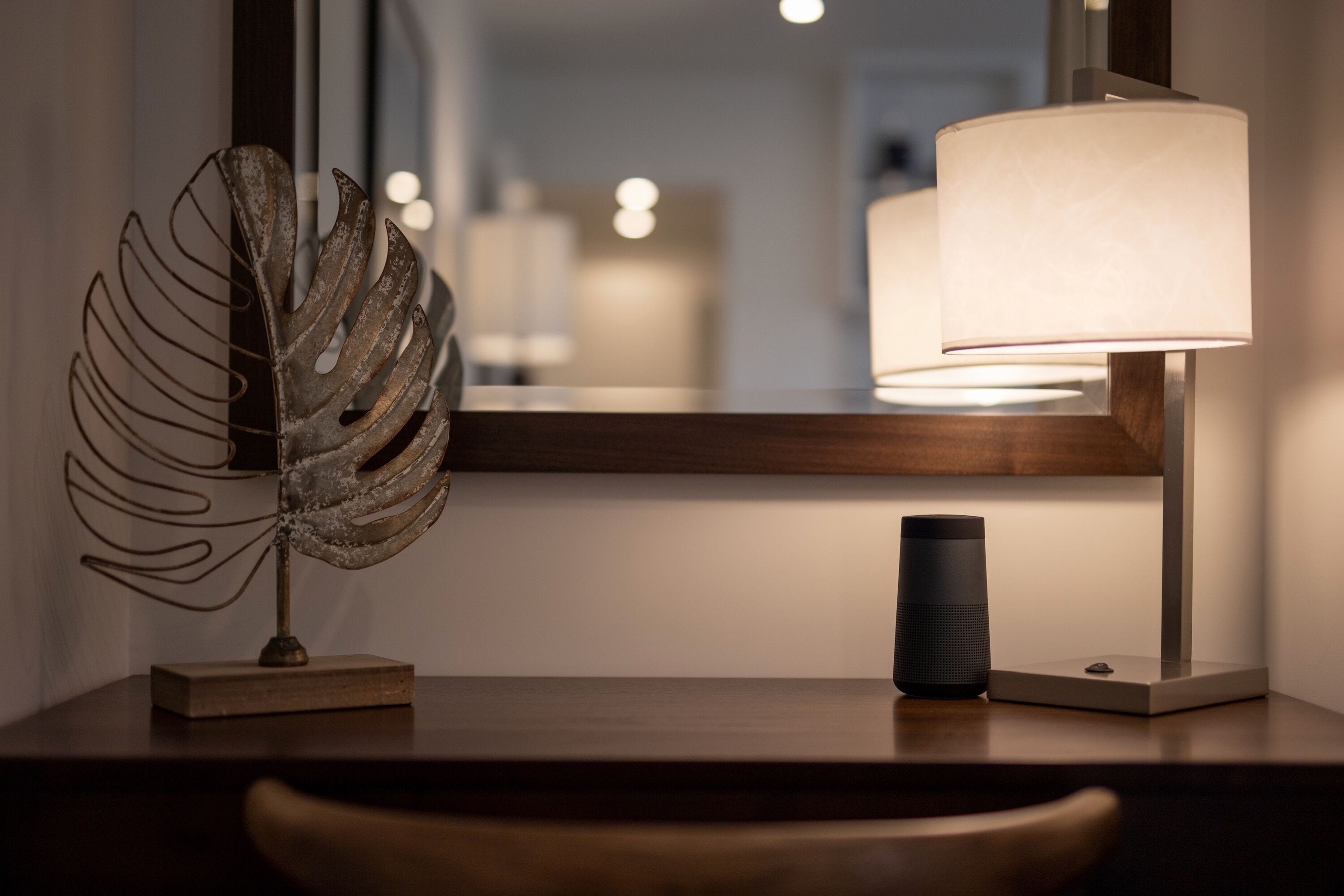 A modern desk with a decorative metal leaf sculpture, a black cylindrical object, and a lamp with a white shade.
