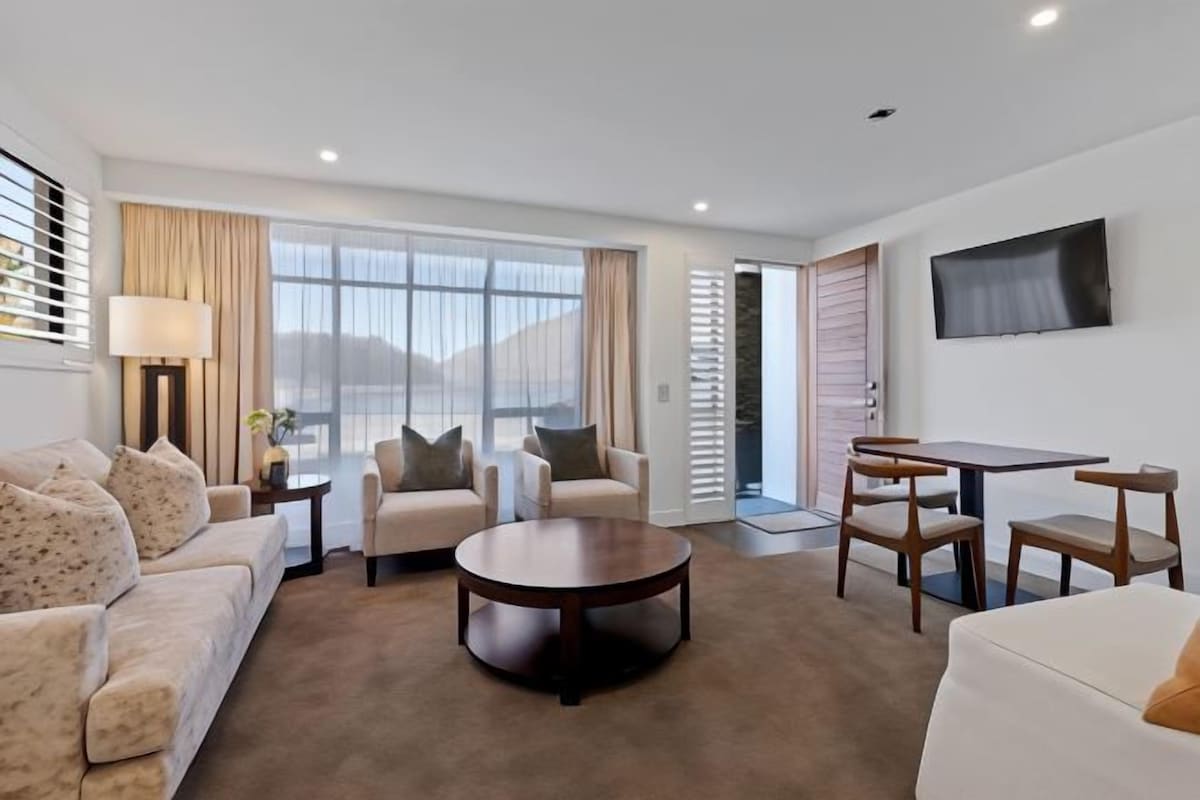 Una habitación de hotel moderna con sofá, dos sillones, una mesa de centro, una mesa de comedor con sillas, televisor montado en la pared y ventanas amplias con cortinas.
