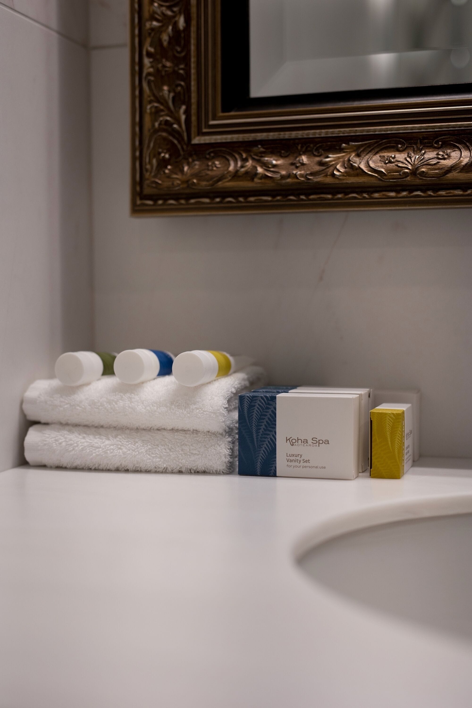 A bathroom vanity with neatly stacked white towels, a bottle of lotion, and a box labeled "Koha Spa Luxury Vanity Set for personal use."