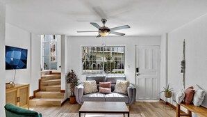 A spacious living room with a grey sofa, wooden coffee table, and a green armchair. A ceiling fan is visible, and there is a staircase leading to another level. - Charming condo walking to beach and town (Tamarindo)