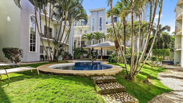 A pool surrounded by a well-maintained lawn, palm trees, and a building with multiple balconies. - Charming condo walking to beach and town (Tamarindo)