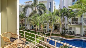 A balcony with wicker furniture, a swimming pool, and a view of a resort with multiple buildings and palm trees. - Charming condo walking to beach and town (Tamarindo)