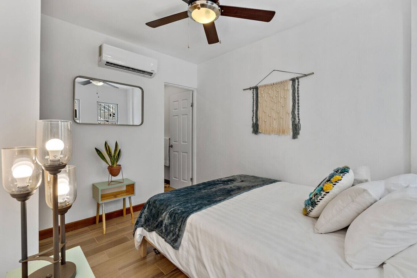 A bedroom with a bed, bedside lamps, a ceiling fan, a mirror, a macrame wall hanging, and a small plant on a wooden nightstand.