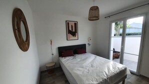 A bedroom with a bed, a round side table, a wall-mounted mirror, a hanging light fixture, and a sliding glass door leading to an outdoor area.