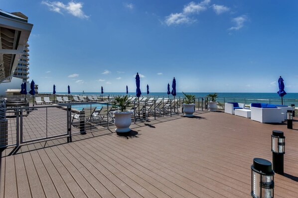 A rooftop pool area with lounge chairs, umbrellas, and a view of the ocean.