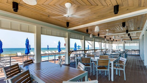 A covered outdoor seating area with wooden tables and chairs, overlooking a pool and beach.