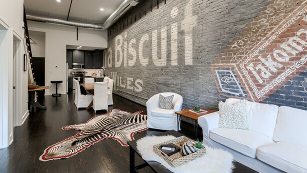 A modern living room with a large brick wall mural, a zebra rug, white furniture, and a kitchen in the background. - New Downtown Luxury Condo, by UNL, stadium & arena, above luxury retail #204 (Lincoln)