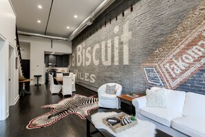 Uma sala de estar moderna com um grande mural de tijolos, um tapete de zebra, móveis brancos e uma cozinha ao fundo.