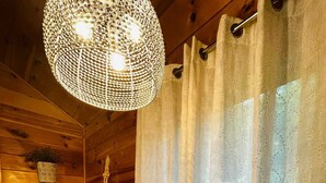 Interior acogedor de una cabaña con piso de madera, un tapete tejido, un plato de ovillos de lana de colores, una lámpara colgante y una cortina.