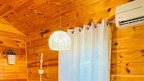Interior de una cabaña de madera con una mesa de comedor, sillas, una lámpara colgante y un ventanal con cortinas.