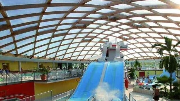 An indoor water slide with a transparent roof structure, surrounded by a pool area with seating and palm trees. - Ruhig Gelegene Fewo 02 - Rustikal und Modern Eingerichtet in Waldnähe (Achslach / Lindenau)