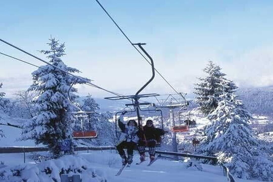 Un télésiège avec deux skieurs assis sur le siège, entouré d’arbres couverts de neige et sous un ciel dégagé.