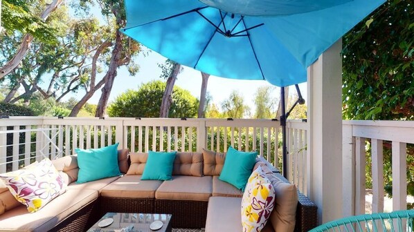 Een overdekt terras met een witte hek, een blauwe parasol en een rieten bank met turquoise kussens.