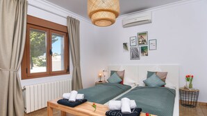 A hotel room with two beds, a wooden bench with towels, a window with curtains, a wall-mounted air conditioner, and framed pictures. - Wonderful and cheerful villa in Javea, Costa Blanca, Spain  with heated pool for 6 persons (Javea)