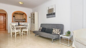 Un salon moderne avec un canapé gris, des chaises blanches, une petite table agrémentée d’objets décoratifs et une cuisine visible à travers une arche.