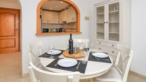Un coin repas avec une table ronde dressée pour quatre personnes, entourée de chaises blanches. Un arc en bois mène à une cuisine dotée de meubles en bois et d’un four à micro-ondes. Un meuble avec des portes en verre est visible sur la droite.