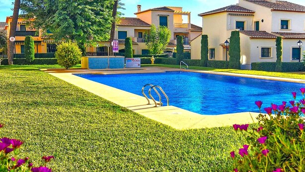 Une piscine entourée de pelouses et de parterres de fleurs bien entretenus, sous un ciel dégagé.
