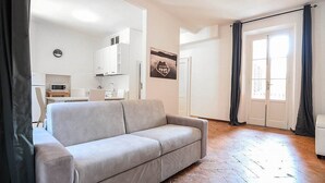 Ein Wohnzimmer mit grauem Sofa, einer Küche im Hintergrund und einem großen Fenster mit Vorhängen.
