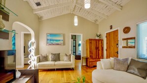 Un salón amplio con techo de madera, un sofá blanco, un sofá redondo blanco, un aparador de madera y un cuadro con una escena de playa.