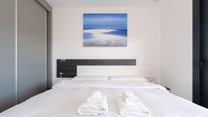 A hotel room with a large bed, white linens, a painting of a beach scene, and a dark headboard.