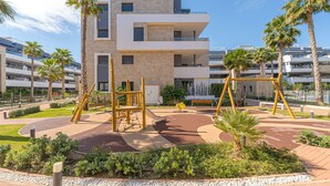 A playground with swings and climbing structures, surrounded by palm trees and a modern building in the background.