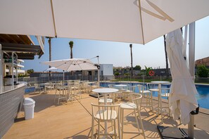 Area piscina all'aperto con sedie e ombrelloni bianchi, una piscina e vista su palme e edifici.