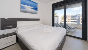 A modern hotel room with a large bed, a bedside table with a lamp, a painting of a beach, and a balcony with chairs.