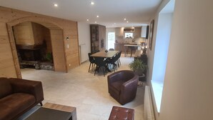 Een moderne woonkamer met een open haard, houten wanden, een eethoek met tafel en stoelen, en een keuken op de achtergrond.