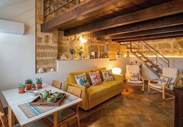 A cozy room with a wooden ceiling, stone walls, a yellow sofa, a dining table with chairs, and a staircase. - Alghero Old Town – Rare Luxury Duplex, Sleeps 6, Steps from the Sea (Alghero)