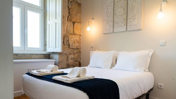 A neatly made bed with white linens and a dark blue blanket, a stone wall, and a window with white shutters. - CASA NINA - Charming apartment with pool on the property (Porto)