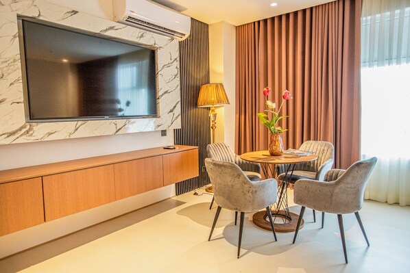 A modern living room with a flat-screen TV mounted on a marble wall, a wooden cabinet, a round dining table with four chairs, and a vase with flowers.