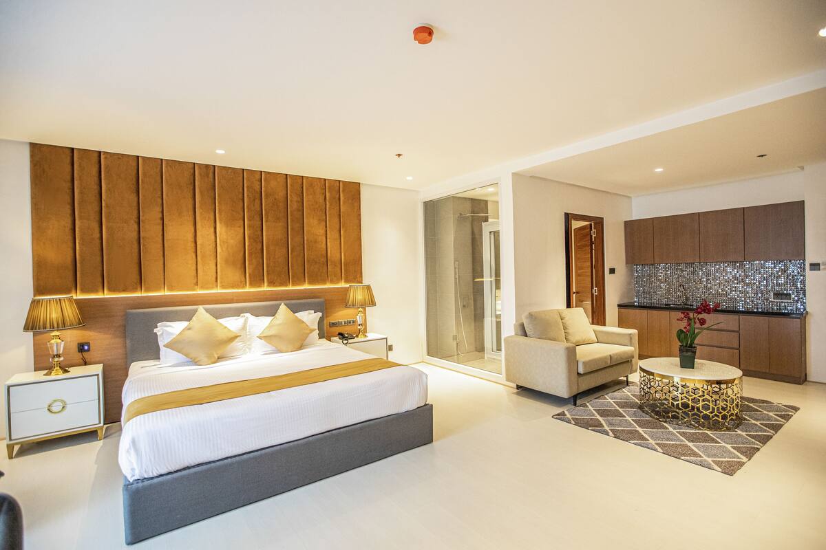 A modern bedroom with a large bed, bedside lamps, a sofa, and a kitchen area visible through an open door.