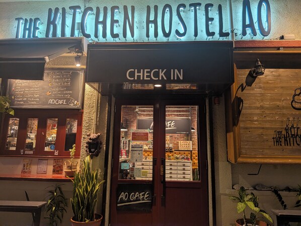 The exterior of The Kitchen Hostel AO with a 'Check In' sign, a menu board, and a 'No Cafe' sign.