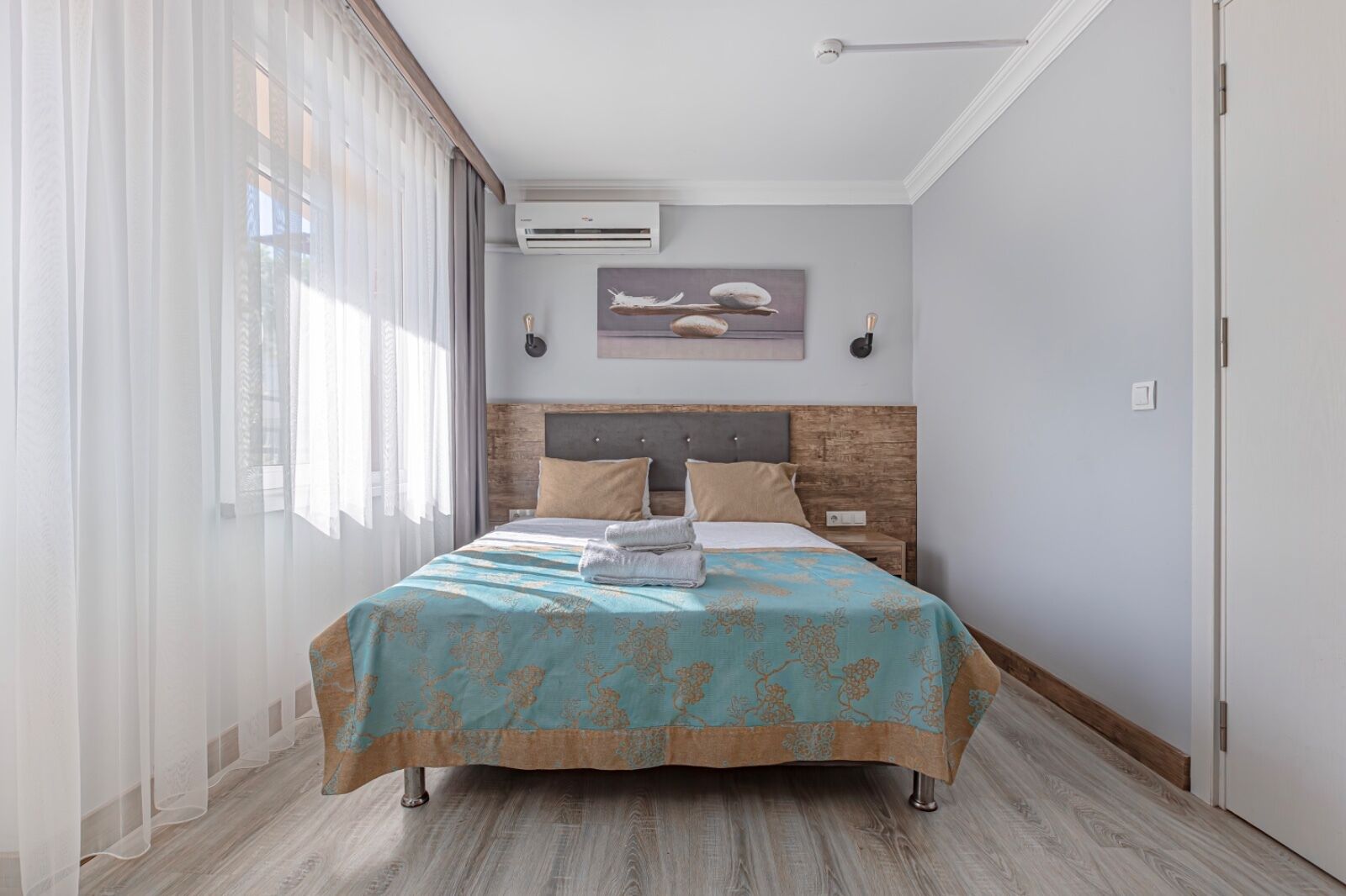 A modern bedroom with a wooden headboard, a bed with a blue and gold patterned bedspread, and a large window with sheer curtains.